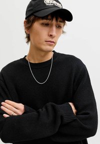 Hombre joven con suéter negro, gorra negra con texto blanco, cadena plateada, pendiente pequeño de aro y anillo, con los brazos cruzados, mirando hacia abajo pensativamente.