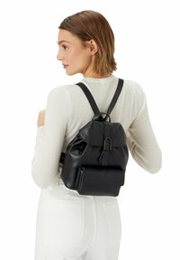 Donna con capelli corti castani che indossa un maglione bianco a coste e pantaloni bianchi, porta uno zaino in pelle nera di piccole dimensioni con un cappuccio a fibbia.