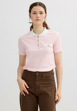 Mujer joven con cabello castaño recogido en una coleta, vistiendo una camiseta polo de manga corta rosa claro con cuello blanco y pantalones marrones de talle alto.