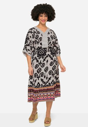 Schwarz und grau gemustertes Midikleid mit V-Ausschnitt, Spitzenakzenten und bunten geometrischen Details am Saum. Die Modells trägt beigefarbene Keilabsätze.