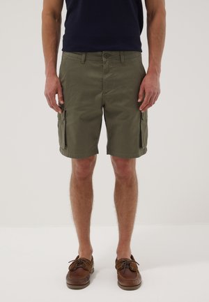 Hombre vestido con pantalones cortos cargo verde oliva, camisa azul marino y zapatos náuticos de cuero marrón, de pie frente a un fondo claro y sencillo.