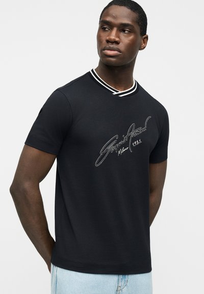 Άνδρας με μαύρο κοντομάνικο πουκάμισο με λευκό κείμενο υπογραφής και ριγέ γιακά, συνδυασμένο με ανοιχτό μπλε τζιν, κοιτάζοντας προς τα πλάγια.