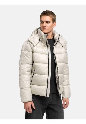Mann trägt eine hellbeige Steppjacke mit hohem Stehkragen und schwarze Hose, schaut seitlich auf einem schlichten hellen Hintergrund.