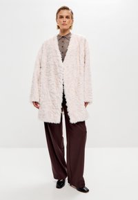 Abrigo largo de piel sintética en un suave tono rosa empolvado, con un ajuste relajado, frente abierto y mangas largas. Se lleva sobre una camisa marrón de botones y pantalones.