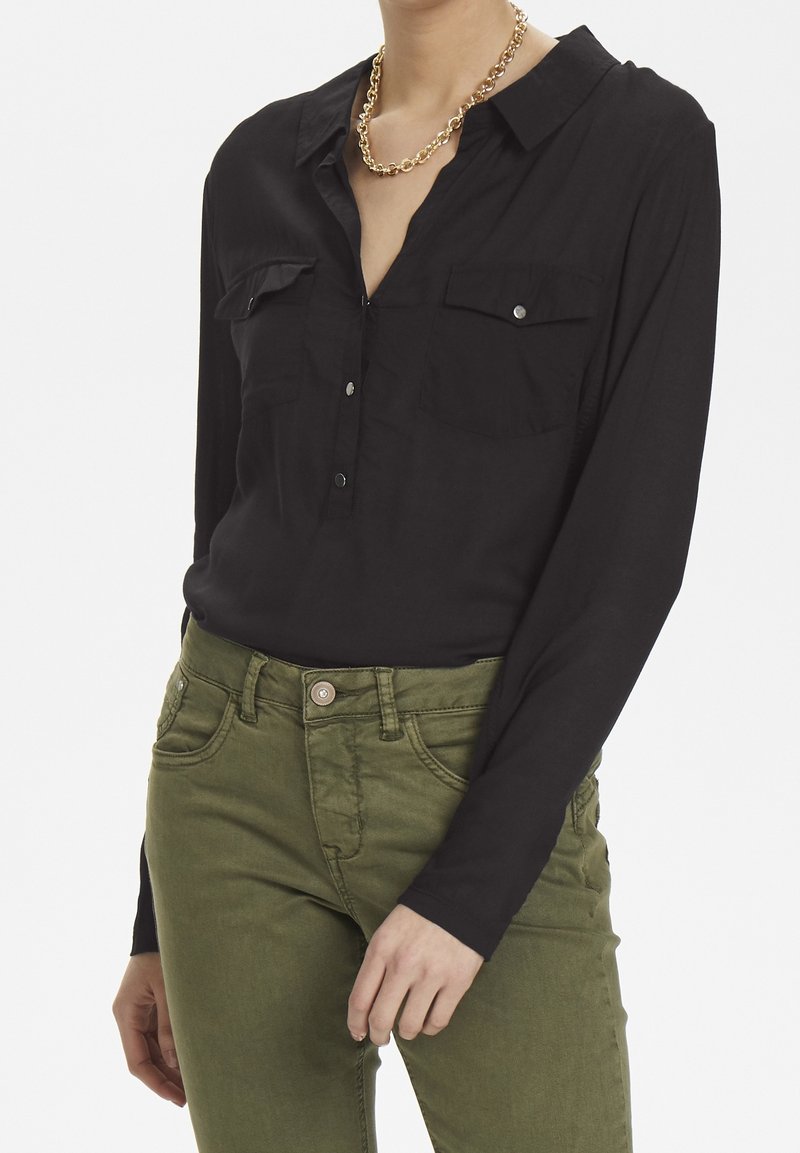 Personne portant une chemise noire à manches longues avec des poches poitrine et un pantalon vert, accessoirisée d'un collier en chaîne dorée.