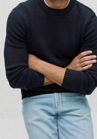Persona che indossa un maglione blu scuro con le maniche arrotolate e jeans azzurri, con le braccia incrociate sul petto su uno sfondo neutro.