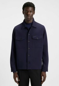 Giacca blu navy con colletto, chiusura frontale con bottoni e due tasche sul petto. Realizzata in tessuto morbido con un'elegante etichetta sul taschino.