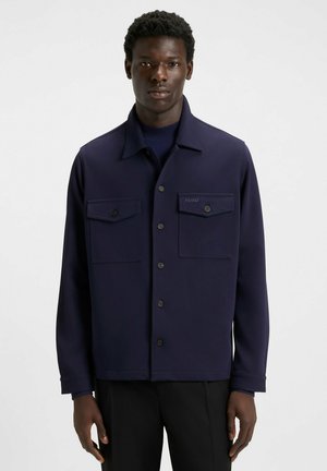 Giacca blu navy con colletto, chiusura frontale con bottoni e due tasche sul petto. Realizzata in tessuto morbido con un'elegante etichetta sul taschino.