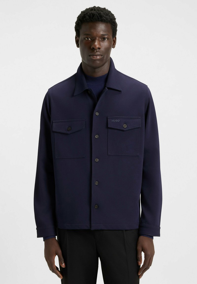 Giacca blu navy con colletto, chiusura frontale con bottoni e due tasche sul petto. Realizzata in tessuto morbido con un'elegante etichetta sul taschino.