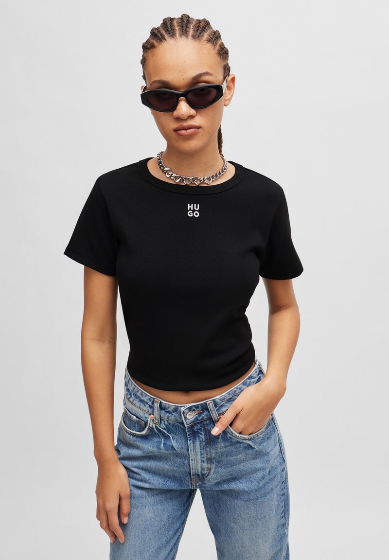 T-shirt noir à manches courtes avec le logo "HUGO" en blanc. Porté avec un jean taille haute bleu clair et un collier en chaîne en argent. Lunettes de soleil noires.