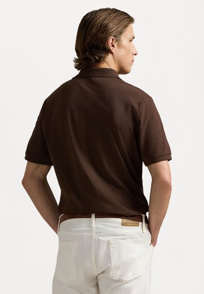 Polo shirt marron à manches courtes avec un col côtelé, associé à un pantalon clair. Le tissu semble lisse et légèrement texturé.