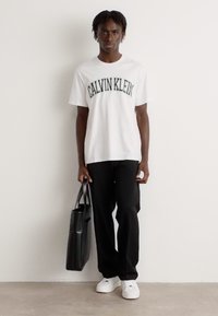 Hvit T-skjorte med kort erm og "CALVIN KLEIN"-logo, svarte vide bukser, hvite joggesko og en svart tote-bag. Ensfarget bakgrunn.