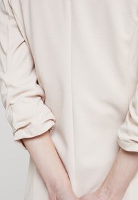 Světle růžový top s texturovanou látkou. Má vyhrnuté rukávy a jednoduchý, volný střih. Viditelné švy podél zad.