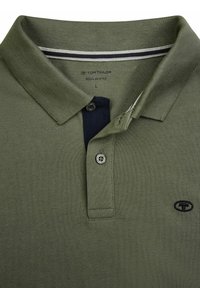 Camiseta polo verde oliva con un tejido texturizado, tres botones y un cuello azul marino. Presenta un pequeño logo bordado en el pecho.