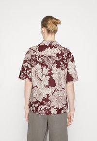 Camicia a maniche corte color bordeaux con motivi floreali color crema, dotata di colletto e vestibilità rilassata. Realizzata in un tessuto leggero.