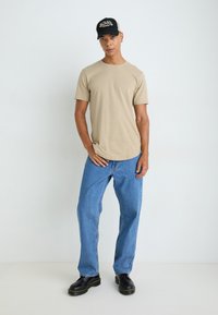 T-shirt beige a girocollo, jeans blu a vestibilità rilassata, stivali neri e un cappellino nero e beige con logo. Design minimalista e aspetto casual.