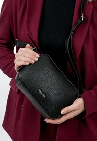 Black leather crossbody bag with a textured finish, rectangular shape, and silver hardware accent, held in hand against a burgundy coat.