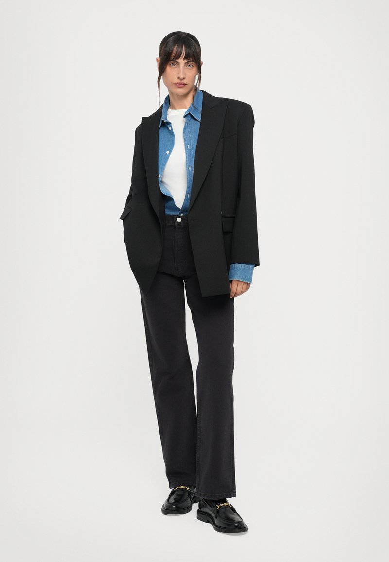 Blazer noir oversize, chemise en denim à boutons, t-shirt blanc, jeans foncés et chaussures noires avec des accents en métal doré.