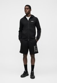 Μαύρο φούτερ με φερμουάρ και ετικέτα λογότυπου, σε συνδυασμό με μαύρο σορτς με λευκή επωνυμία. Μαύρα αθλητικά παπούτσια και κάλτσες μέχρι τον αστράγαλο ολοκληρώνουν την εμφάνιση.