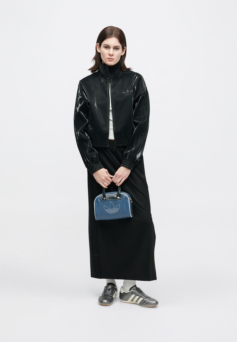 Frau in schwarzer, glänzender Zip-Jacke und langem schwarzen Rock, die eine blaue Adidas-Handtasche hält und graue Adidas-Sneaker mit weißen Socken trägt.