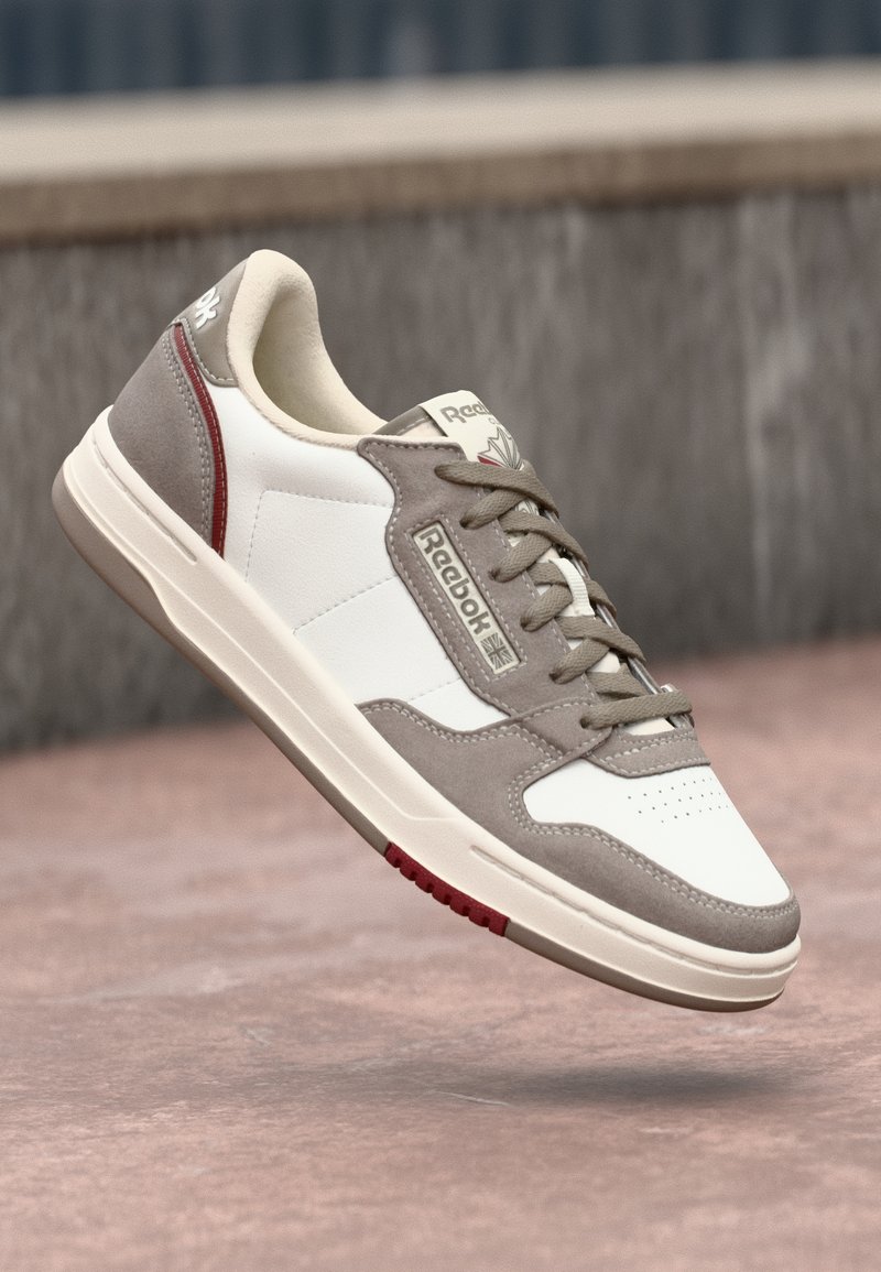 Sneakers albe și gri cu un finisaj din piele întoarsă texturată și piele, având o zonă a degetelor perforată, șireturi gri și un accent roșu pe talpă.