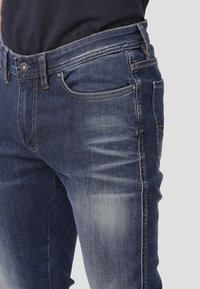 Jeans de mezclilla en azul oscuro con una textura ligeramente desgastada, que cuentan con cinco bolsillos, un cierre de botón y costuras visibles a lo largo de las costuras.