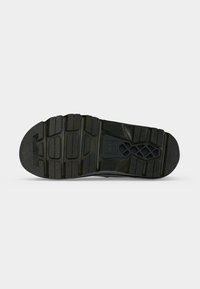 Semelle de chaussure noire avec des motifs de bande de roulement épais et des zones de grip texturées, vue de dessous sur un fond clair.