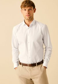 Weißes Hemd mit langen Ärmeln, aus glattem Material gefertigt. Getragen mit beigen Hosen und einem braunen Gürtel. Schlichtes und klassisches Design.