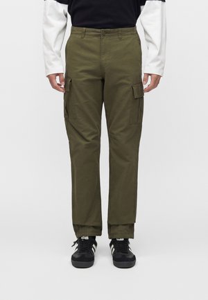 Man wearing olive green cargo pants with side pockets, black and white sneakers, and a black and white long-sleeve shirt, standing on a white background.