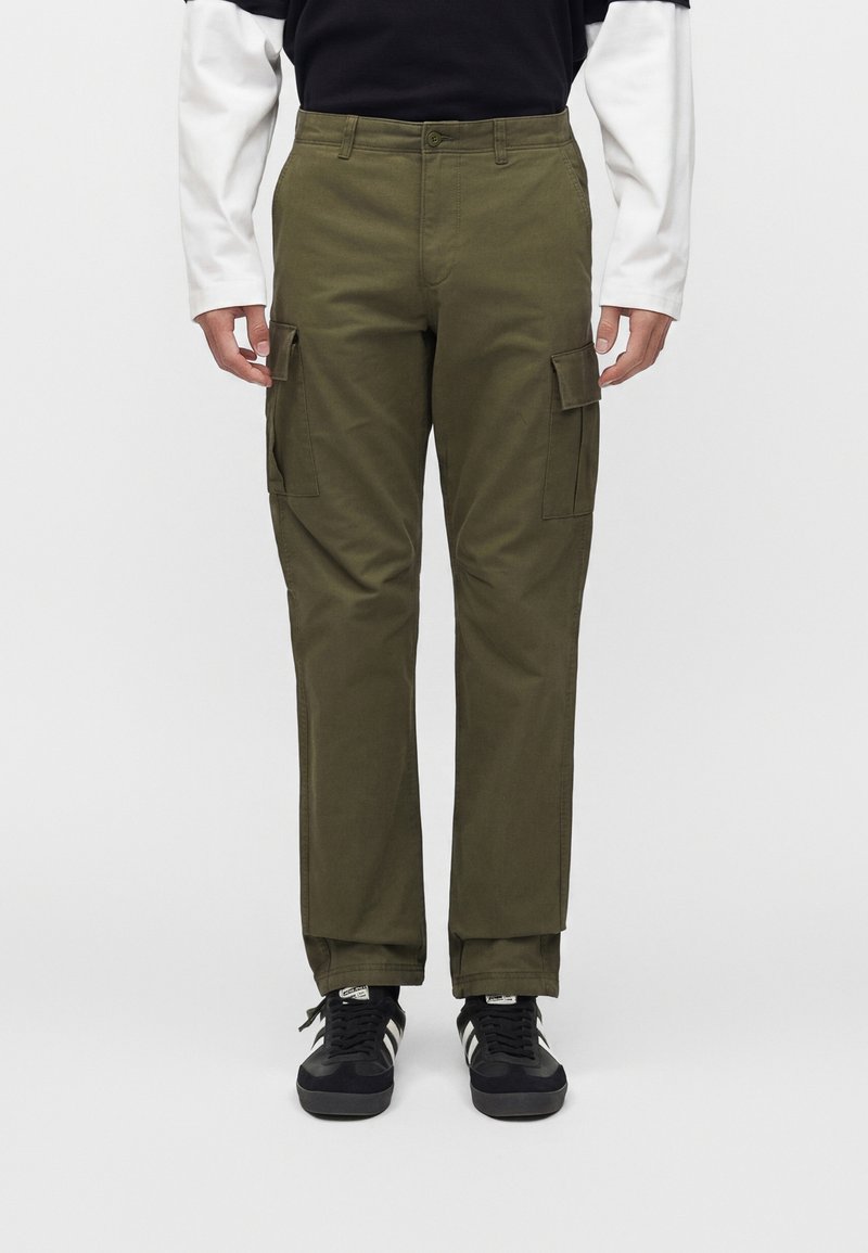 Uomo con pantaloni cargo verde oliva con tasche laterali, scarpe da ginnastica nere e bianche e una maglia a maniche lunghe bianca e nera, in piedi su sfondo bianco.