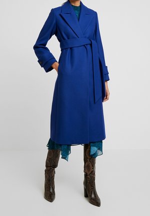 Kvinde iført en knælængde kongeblå frakke med bælte over en grønblå kjole med flæsekant, kombineret med knæhøje brune slangeskinsstøvler.