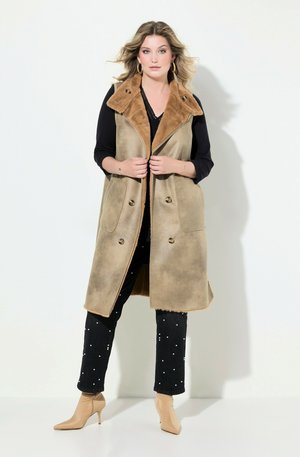 Bruine nepleer schapenvachtvest met een brede kraag, knopenfront en twee grote zakken, gelaagd over een zwarte top en jeans met witte accenten.