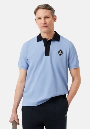 Hombre con polo azul claro con cuello y logo negros, pantalones negros, sosteniendo un objeto marrón en su mano izquierda, de pie contra un fondo blanco.