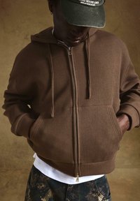 Rjava rebrasta zip-up hoodie s kapuco, sprednjima žepoma in belim majico pod njim. Temno vzorčaste hlače dopolnjujejo sproščen videz.