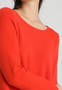 Roter Pullover mit rundem Halsausschnitt und langen Ärmeln, aus weichem Stoff mit glatter Textur und minimalen Nähdetails gefertigt.