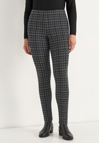 Nainen, jolla on päällään musta-harmaa ruutukuosiset slim fit -housut, mustat korkeat nilkkasaappaat ja musta pitkähihais top.