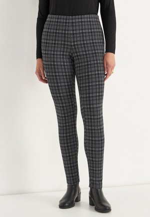 Frau in schwarz-weißen karierten Slim-Fit-Hosen, mit schwarzen, knöchelhohen Stiefeln mit Absatz und einem schwarzen Langarmoberteil.