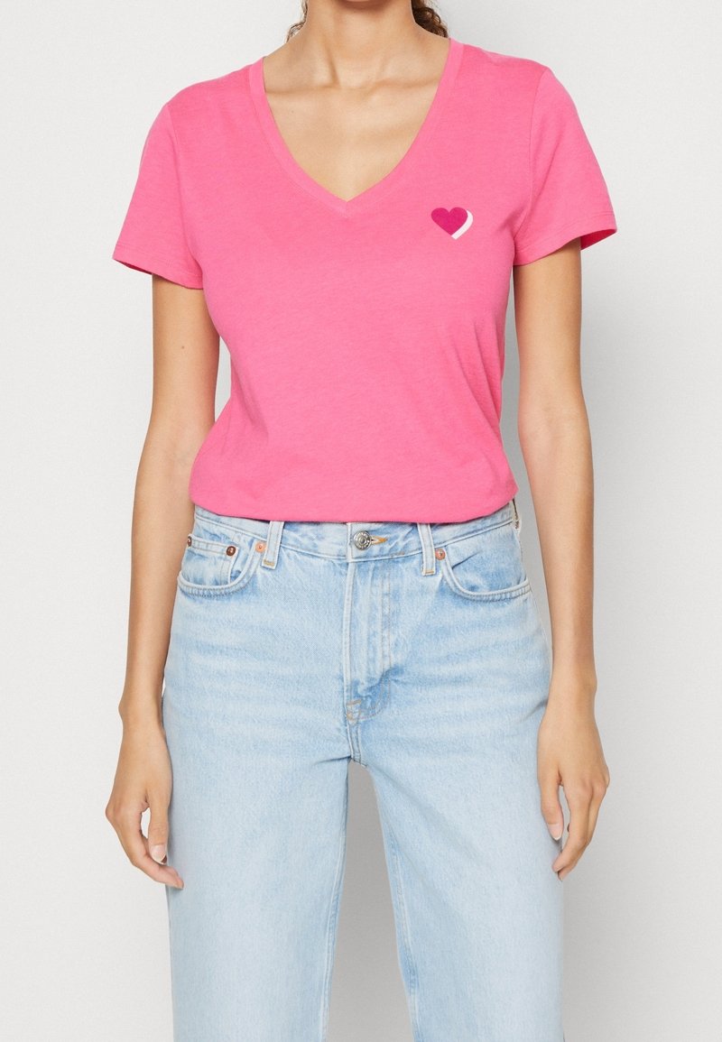 T-shirt rose en V en tissu doux, arborant un petit graphique en forme de cœur en rose foncé et clair sur la poitrine gauche, associé à un jean bleu clair.