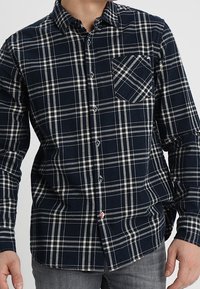 Camicia a quadri blu navy e crema con colletto, maniche lunghe e tasca sul petto con un motivo a righe diagonali.