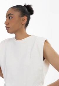Luźna koszulka w kolorze kremowym z okrągłym dekoltem, szerokimi otworami na ramiona oraz gładką bawełnianą strukturą, prezentująca minimalistyczny design.