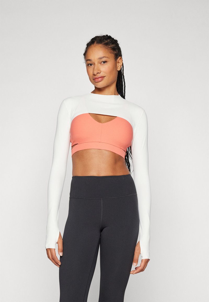 Dugih rukava crop top u bijeloj i koraljnoj boji, s dizajnom rezanja. Uparen s crnim tajicama visoko struka, koje pokazuju glatku teksturu.