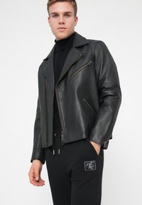 Črna usnjena motoristična jakna s srebrnimi zadrgami, z asimetričnim sprednjim delom, širokimi reverji in gladko teksturo. Nosi se čez črno turtleneck majico.
