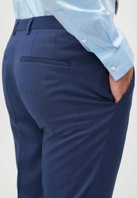 Άνδρας που φοράει κομμένα ναυτικά μπλε παντελόνια και ανοιχτό μπλε πουκάμισο με το χέρι στην τσέπη, φαίνεται από τη μέση και κάτω.