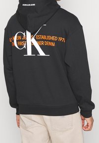 Černá mikina s kapucí s bílými a oranžovými textovými grafikami, včetně nápisu "CALVIN KLEIN JEANS" a "CK", s měkkou texturou a žebrovaným lemem.