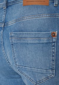 Jasnoniebieskie dżinsy z widoczną tylną kieszenią oraz brązową skórzaną łatą z wytłoczonym tekstem. Szwy w kontrastowym kolorze.