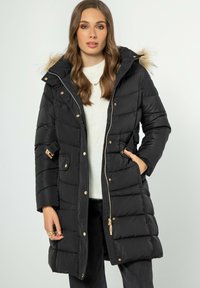 Abrigo de plumas negro con capucha forrada de piel sintética desmontable, detalles en dorado, cremallera, bolsillos laterales y textura acolchada. Combinado con un suéter ligero.