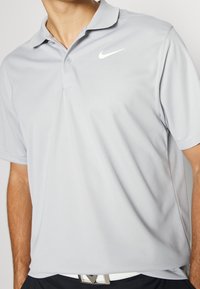 Světle šedé polo tričko vyrobené z hladké, prodyšné tkaniny. Obsahuje klasický límec, zapínání na knoflíky a bílé logo Nike na hrudi.