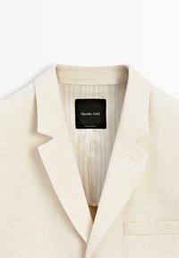 Beżowa marynarka o teksturowanej powierzchni z klapą z wycięciem, kieszenią na piersi i paskowanym podszewką wewnętrzną, z czarną metką wewnątrz kołnierza.