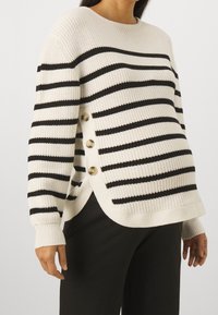 Striped sweater in cream and black, featuring ribbed texture, rounded neckline, and side buttons. Designed with a relaxed fit and split hem.