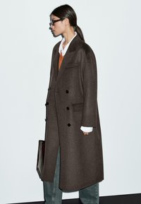 Manteau en laine marron avec un design croisé, de grands boutons noirs et deux poches latérales. Porté sur un pull orange et une chemise blanche.